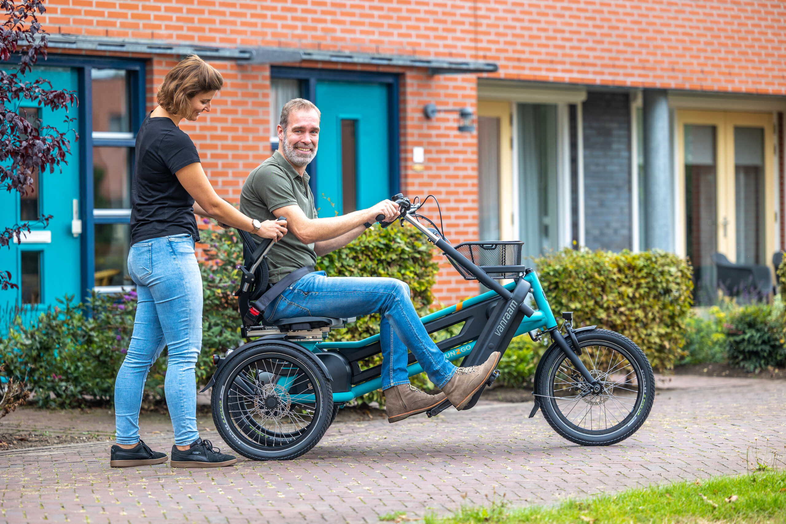 Un homme souriant sur un vélo adapté, aidé par une femme, dans un cadre résidentiel.