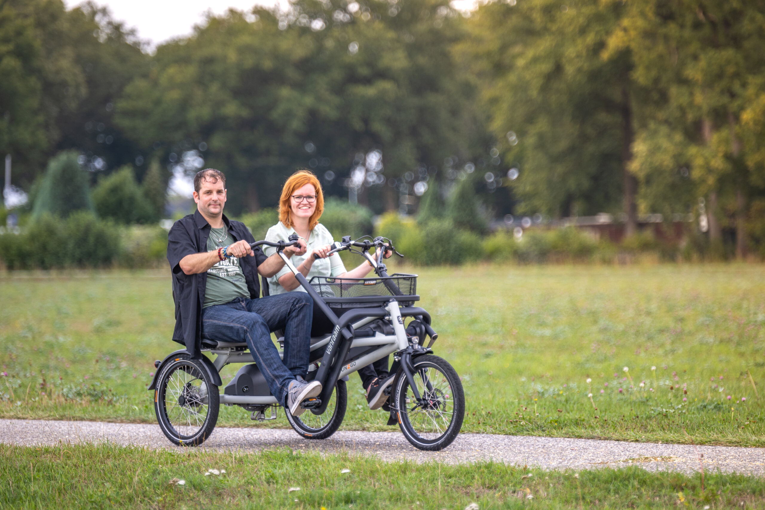 Couple souriant sur un vélo à deux places, roulant sur un chemin verdoyant en Normandie. Location de vélos pour balades.