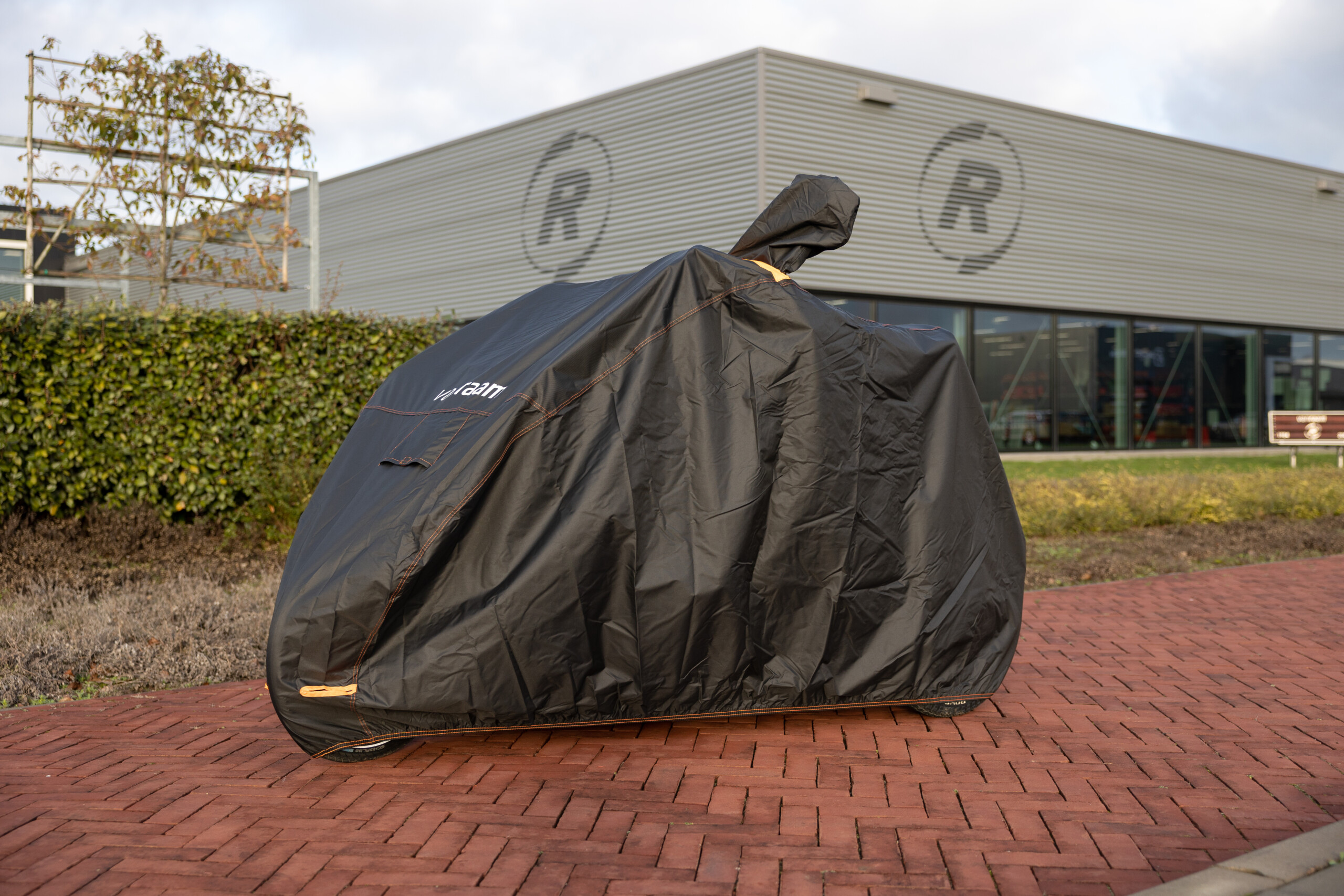 Vélo recouvert d'une housse noire, stationné sur une allée en pavés, devant un bâtiment moderne.