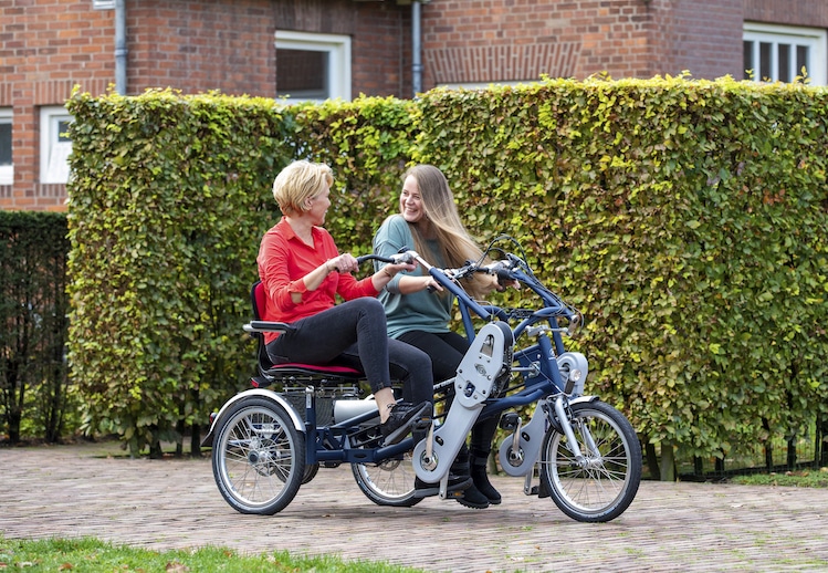 Deux femmes souriantes sur un vélo à deux places, devant une haie, prêtes pour une balade en Normandie.