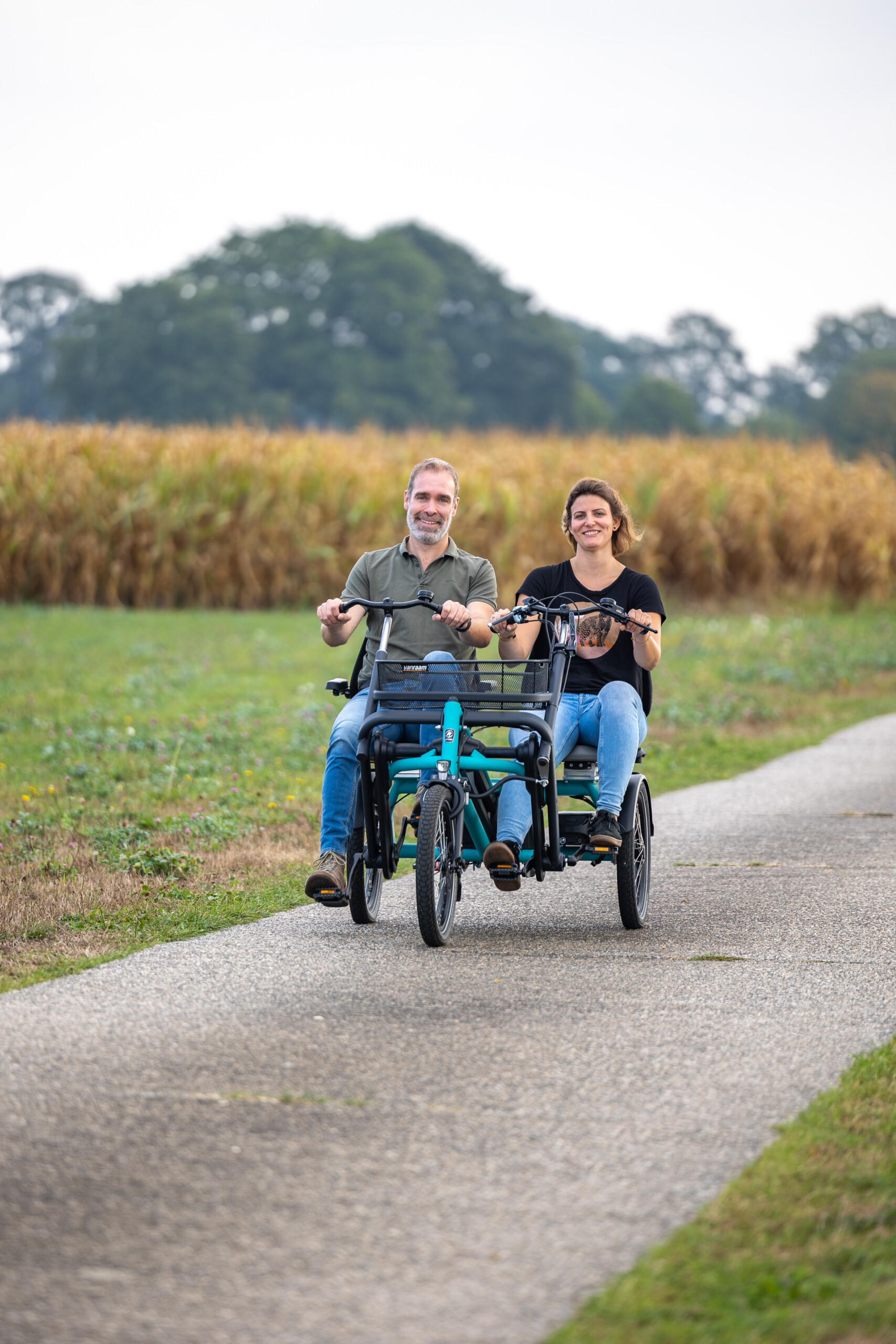 Deux personnes souriantes, sur des vélos côte à côte, avancent sur une route en pleine nature.