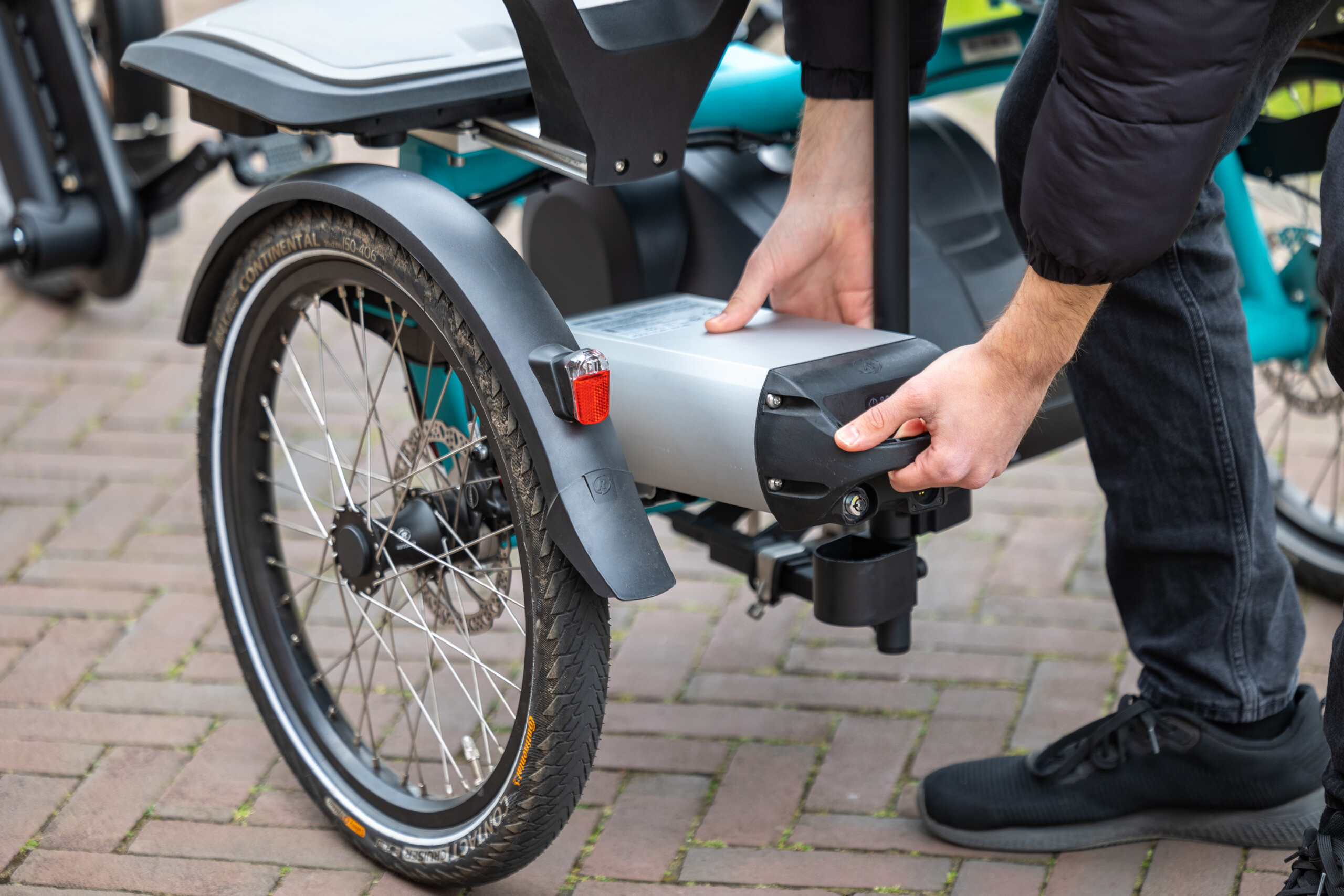 Main d'un utilisateur fixant une batterie à l'arrière d'un vélo électrique sur un sol pavé.