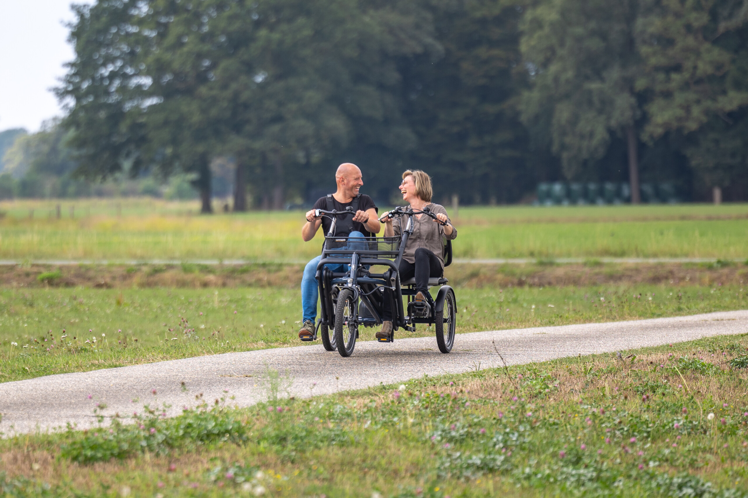 Deux personnes rient ensemble sur un vélo à trois roues, sur un chemin en plein air, entouré de verdure.