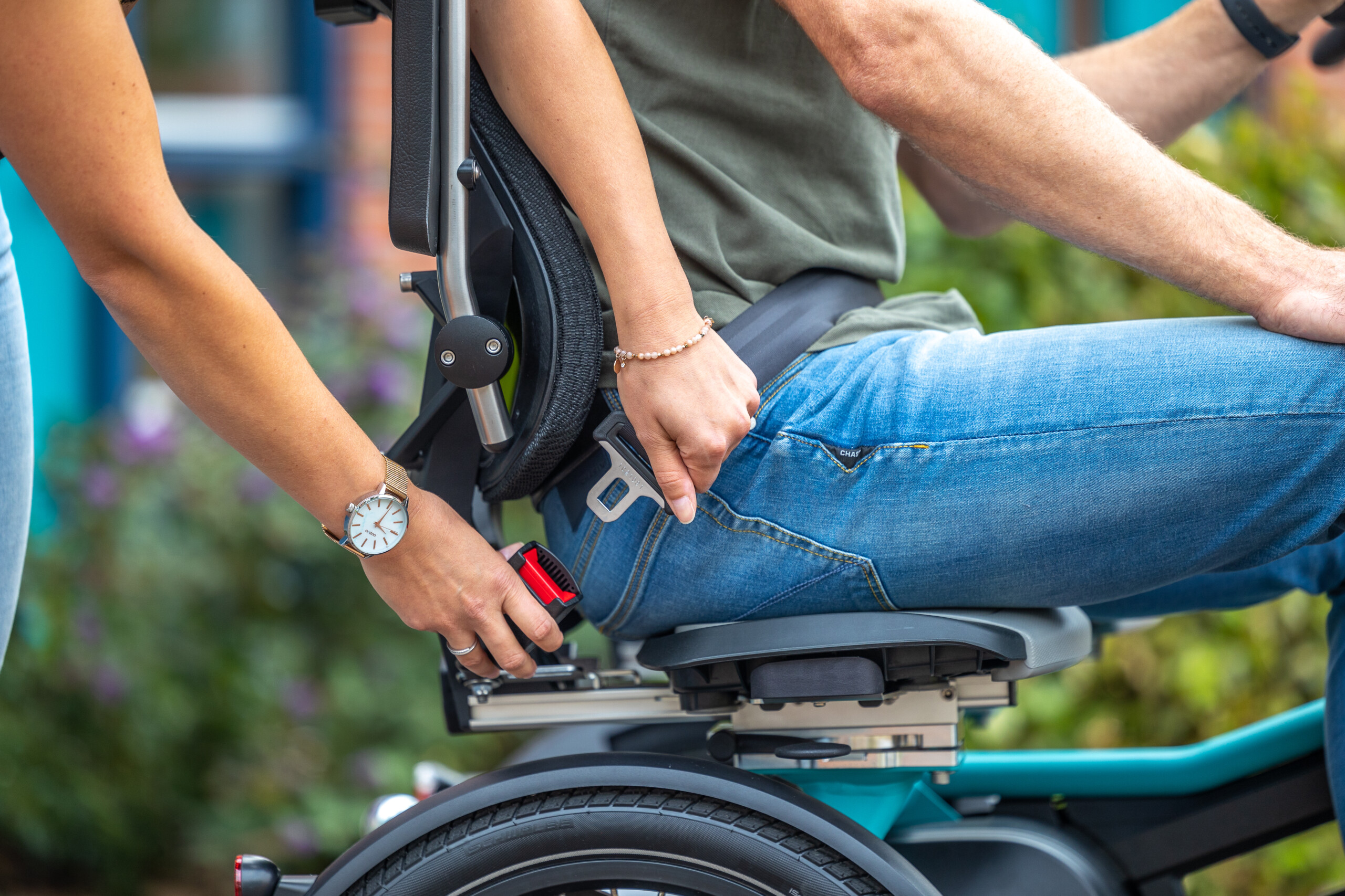 Une personne aide une autre à attacher sa ceinture sur un vélo adapté, dans un cadre extérieur.
