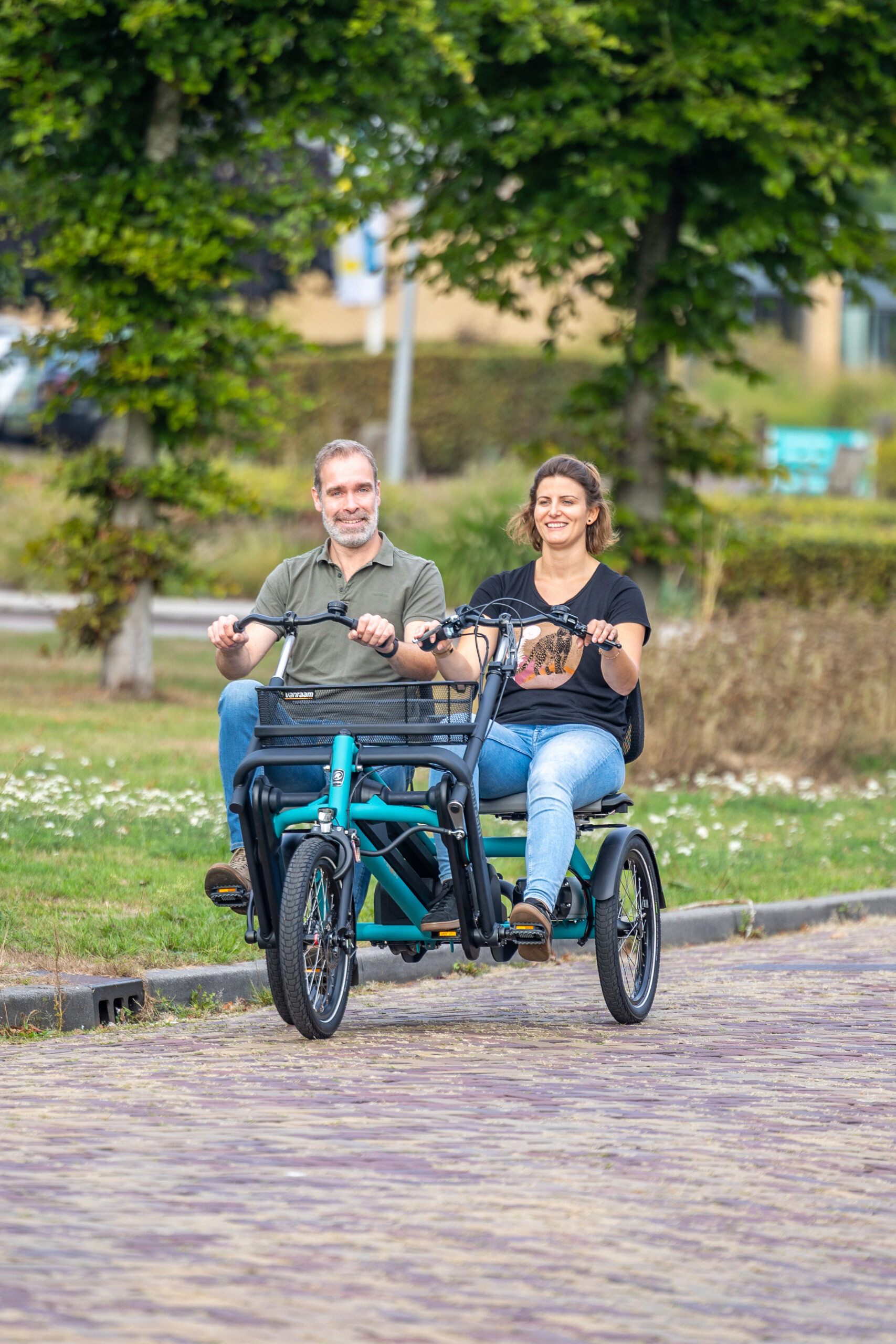 Deux personnes souriantes sur un vélo adapté, roulant sur une voie pavée, entourées de verdure.