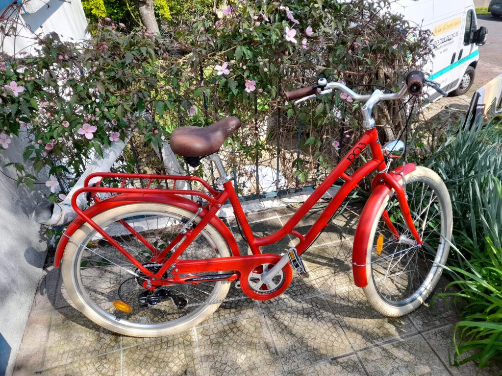 Vélo rouge stationné sur un dallage, entouré de fleurs, prêt à être loué pour des balades en Normandie.