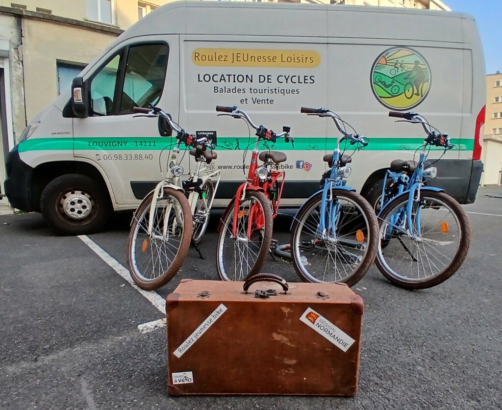 4 Vélos 1903 d'ARCADE CYCLES en location devant le véhicule "Roulez JEUnesse Loisirs" pour balades en Normandie. Une valise vintage est posée au sol au premier plan pour symboliser les vacances et le voyage.