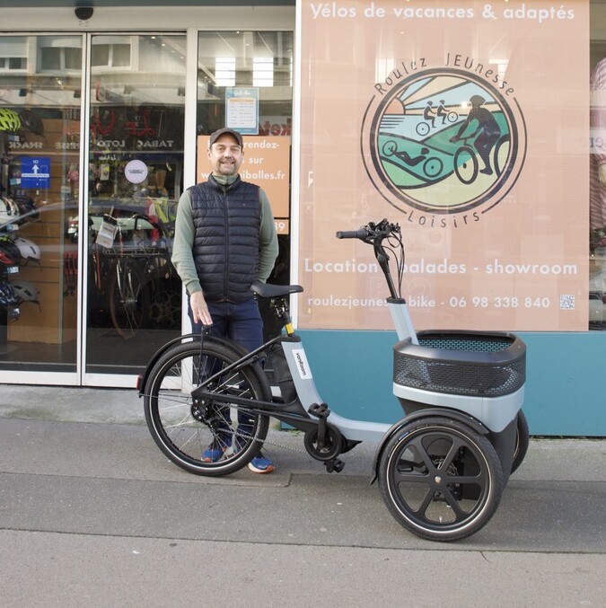 Personne devant un magasin de location de vélos "Roulez JEUnesse Loisirs" à Caen, avec un vélo cargo.