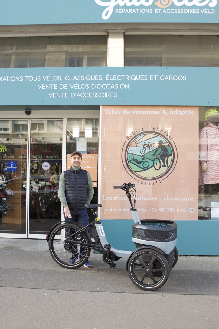 Personne devant un magasin de location de vélos "Roulez JEUnesse Loisirs" à Caen, avec un vélo cargo Thuja pour personnes âgées.